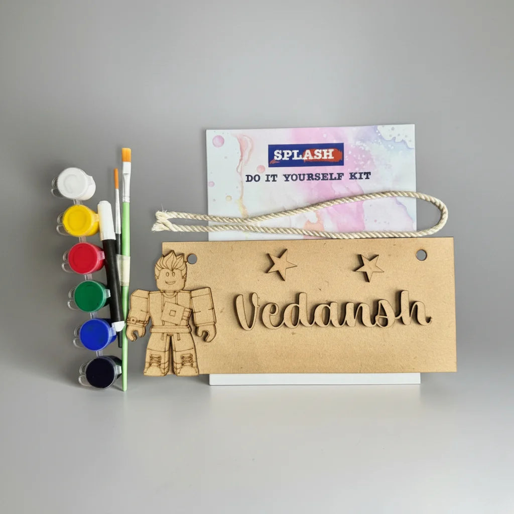 A DIY craft kit labeled 'SPLASH DO IT YOURSELF KIT' with a wooden astronaut figure, a wooden sign reading 'Vedansh', a rope handle, and paint pots with paintbrushes on a light gray background.