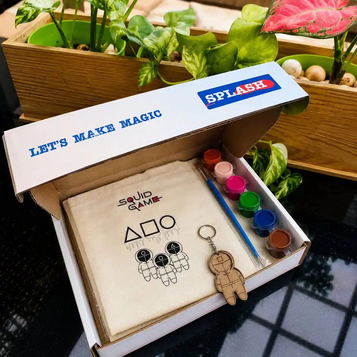 An open white gift box labeled 'LET'S MAKE MAGIC' and 'SPLASH' contains a beige fabric art kit with the 'Squid Game' logo, a small wooden astronaut keychain, and a set of paint pots and a paintbrush. The box is styled on a dark reflective surface with green potted plants in wooden boxes in the background.