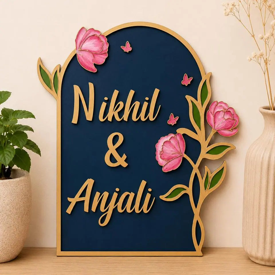 Decorative wall art with 'Nikhil & Anjali' on a blue background with pink flowers and gold frame.