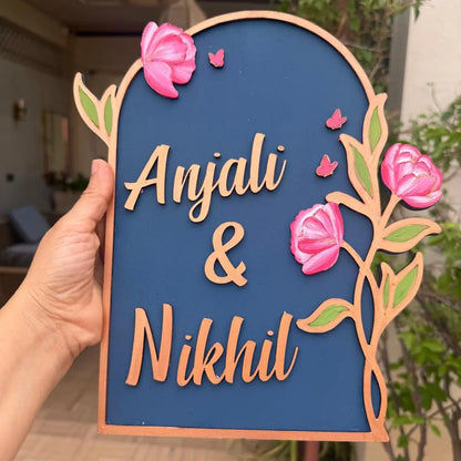 Decorative sign with 'Anjali & Nikhil' surrounded by pink flowers and green leaves.
