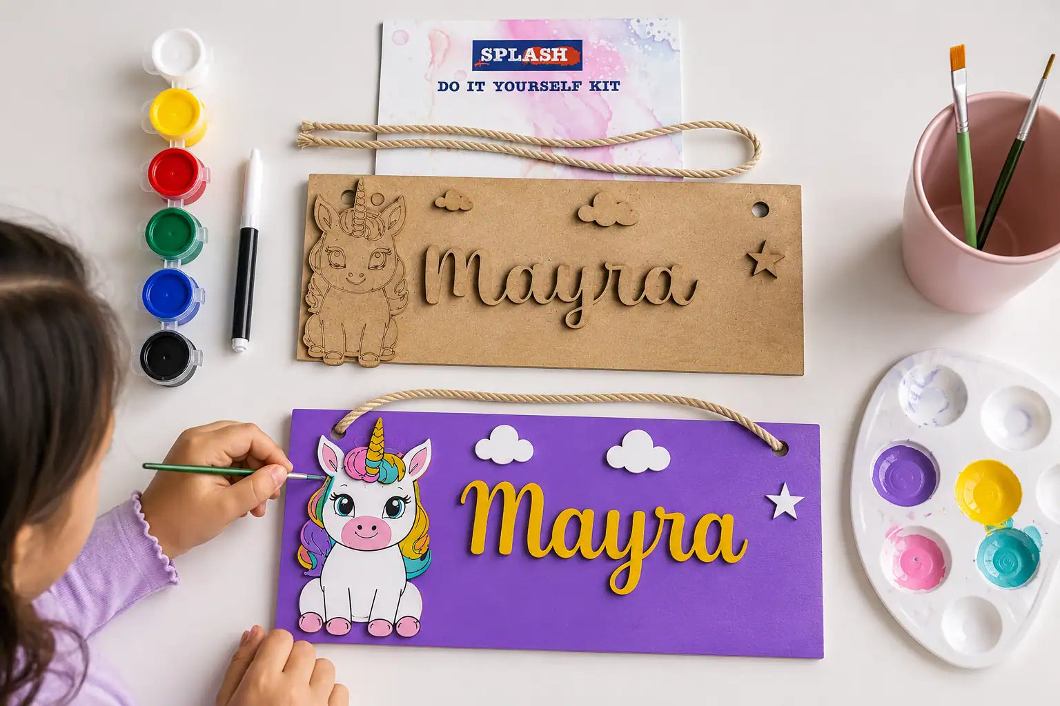 Child painting a unicorn-themed nameplate with 'Mayra' on a table with art supplies.